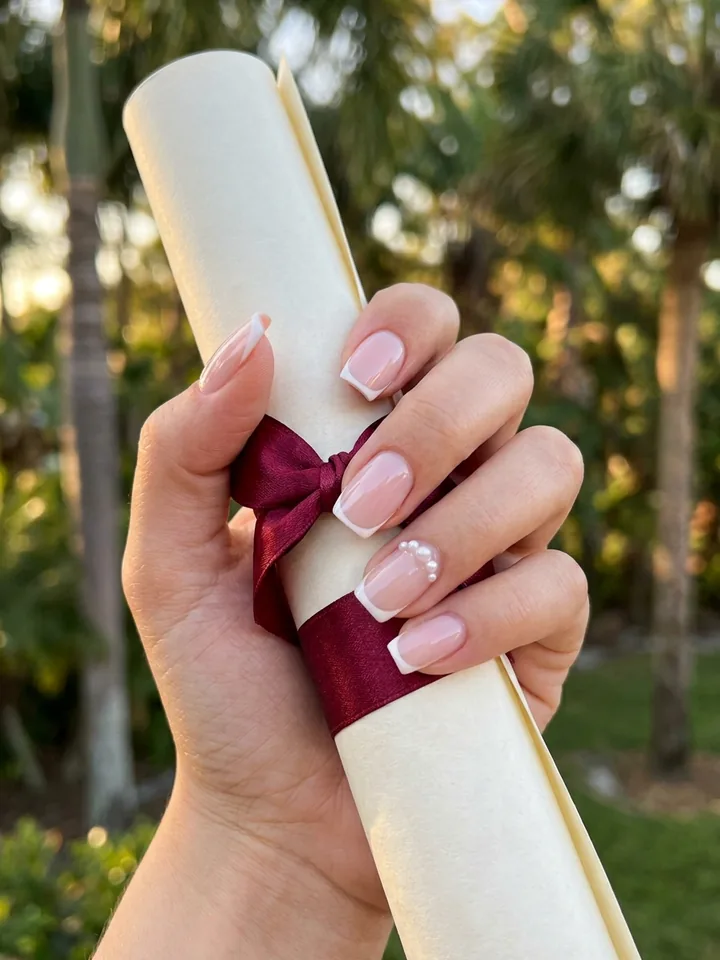 Milky pink French tip nails with pearl accents holding a diploma with a burgundy ribbon